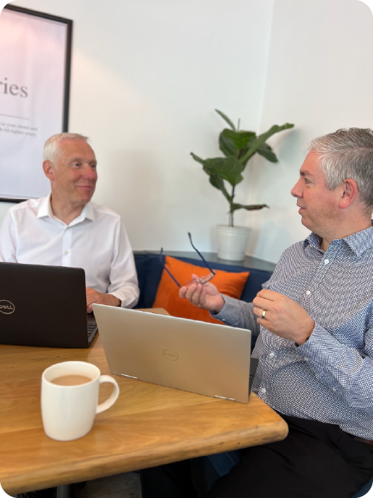Two men sitting at a table with laptops, engaged in a conversation, discussing business IT solutions.