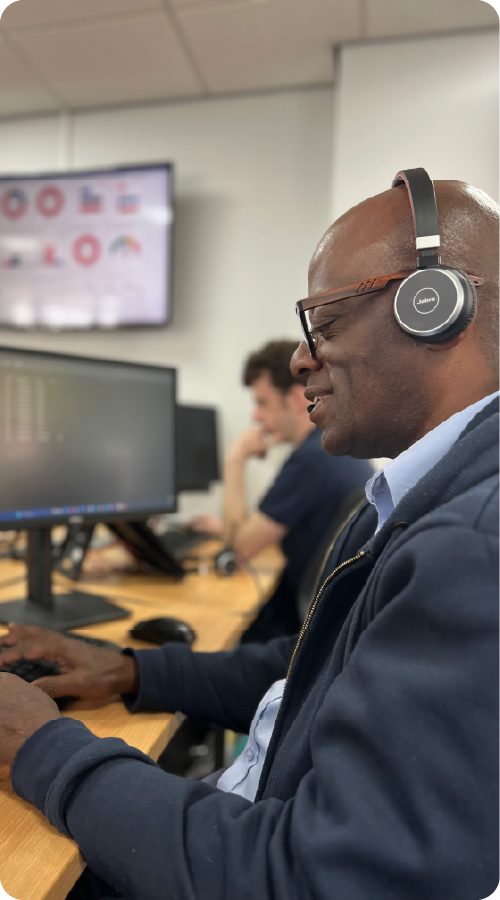 Man wearing headphones sitting at a desk with multiple computer monitors in an office setting providing IT support to clients.