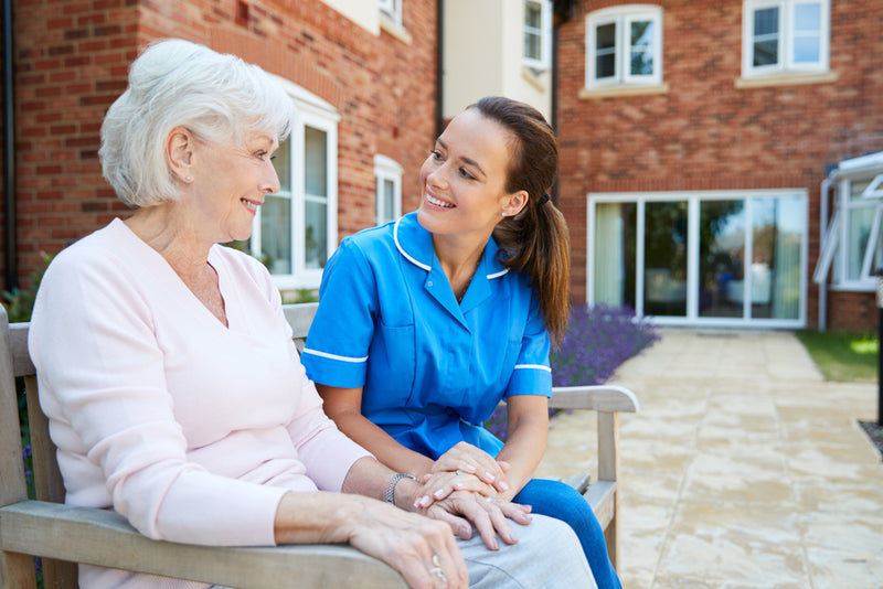 Nurse in blue uniform sitting with an elderly woman on a bench outside a building.