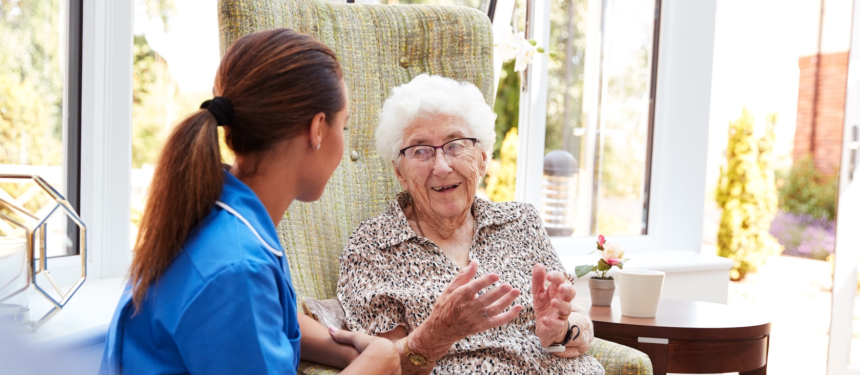 Senior Woman Sitting In Chair And Talking With Nurse In Retirement Home