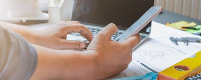Using a laptop and phone at a desk with hard hats, spirit-level and paper plans on the desk.